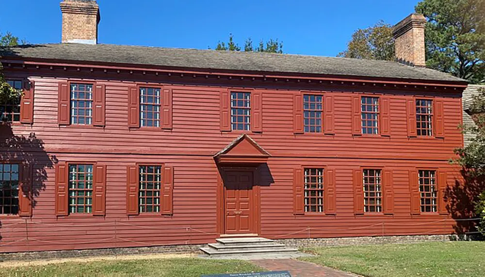 This image shows a two-story red colonial house with green shutters a central doorway with a small portico and a chimney against a clear blue sky