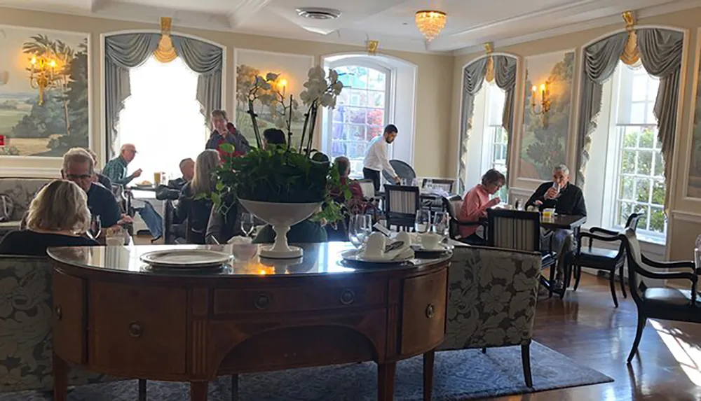 A group of people is dining in an elegantly decorated room with natural light pouring in from large windows