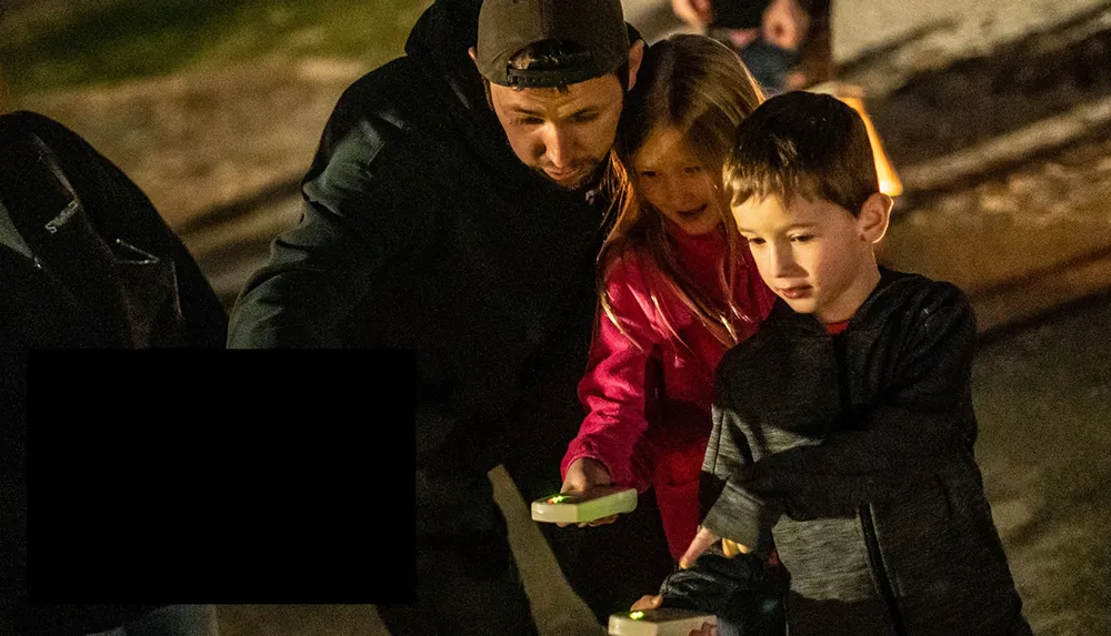 An adult and two children are engaged in an activity at night holding glowing devices with a look of concentration and curiosity