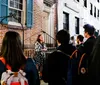 A group of people is attentively listening to a woman who appears to be giving a tour or a talk at night in front of a building with brick walls and blue shutters