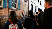 A group of people is attentively listening to a woman who appears to be giving a tour or a talk at night in front of a building with brick walls and blue shutters.