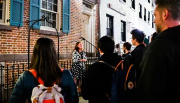 A group of people is attentively listening to a woman who appears to be giving a tour or a talk at night in front of a building with brick walls and blue shutters.