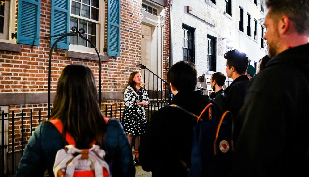 A group of people is attentively listening to a woman who appears to be giving a tour or a talk at night in front of a building with brick walls and blue shutters