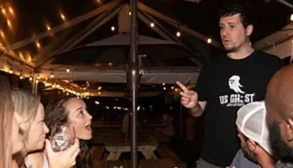 A man appears to be speaking or explaining something to an attentive group of people at an outdoor evening event adorned with string lights