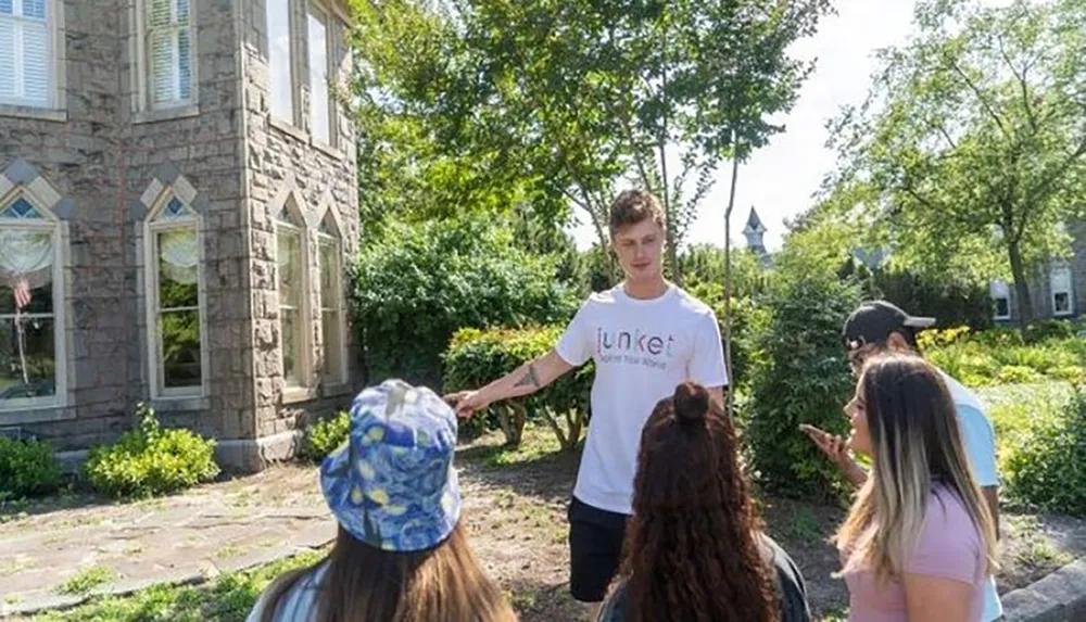 A young man wearing a shirt with a logo is speaking to a group of attentive individuals outdoors near a stone building with greenery around