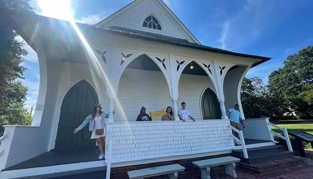 A group of people is standing on the steps of a picturesque white church on a sunny day with sun rays beaming down on them