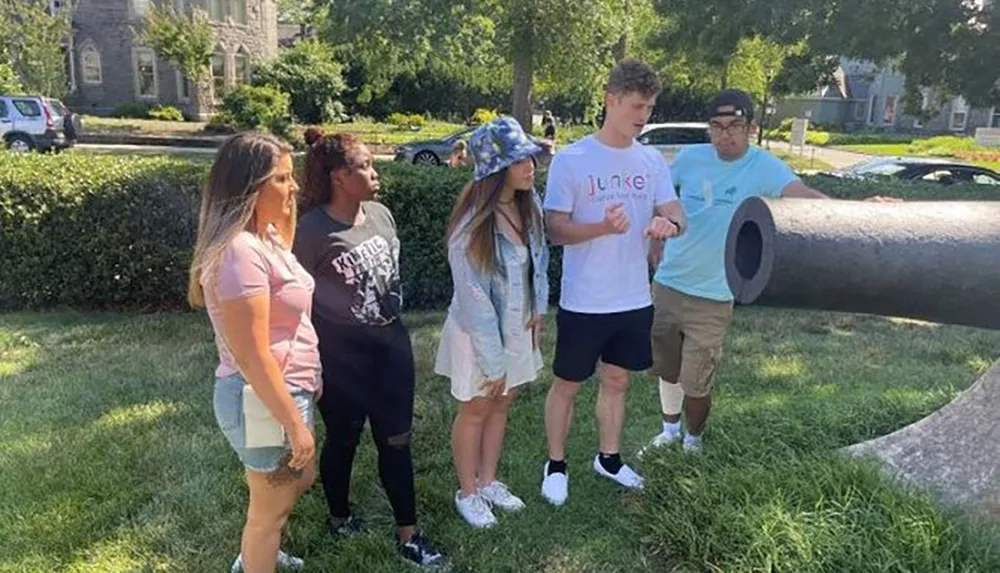 A group of five individuals are standing outdoors during the daytime with puzzled expressions possibly contemplating a large cylindrical object on the ground to their right