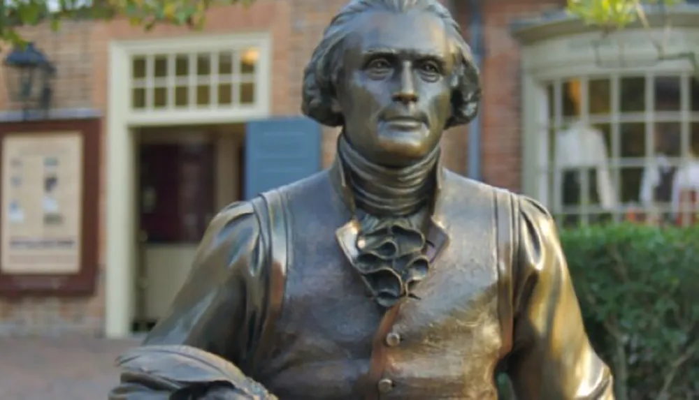 The image shows a bronze statue of a historical figure standing in front of an old brick building with a colonial design