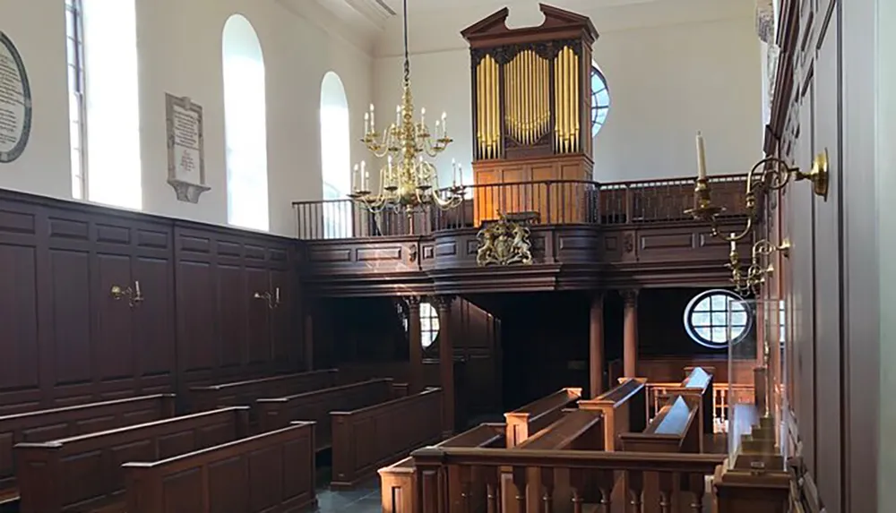 The image shows the interior of a classic church featuring wooden pews a balcony with an organ and ornate chandeliers bathed in natural light from arched windows