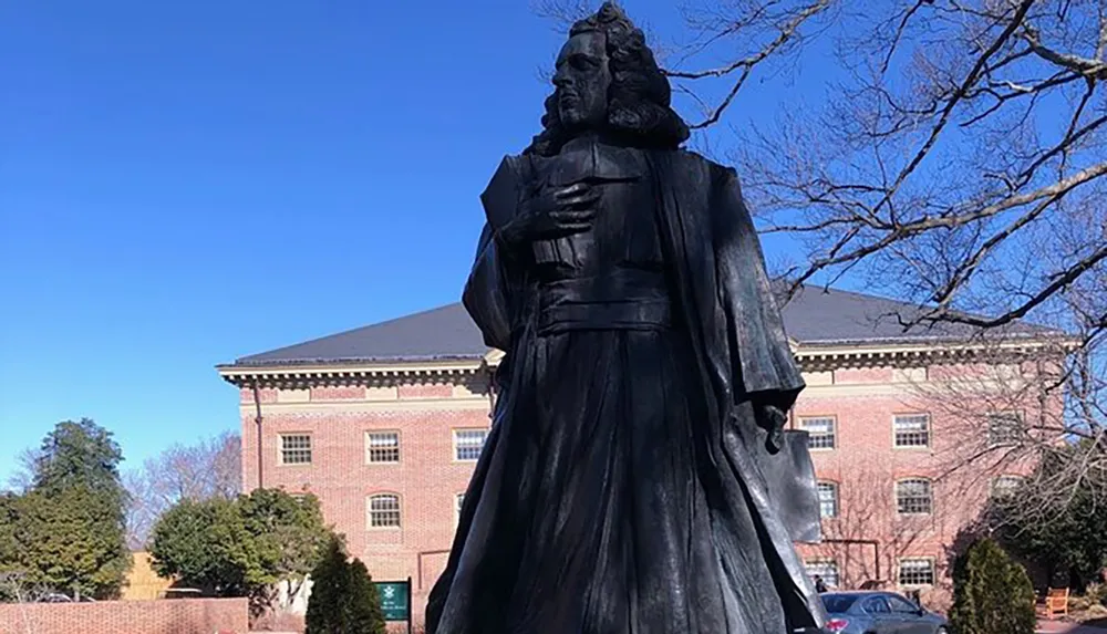 The image shows a bronze statue of a historical figure with long hair and 17th or 18th-century clothing standing in front of a red brick building under a clear blue sky