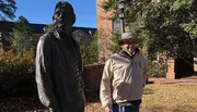 A person is standing and smiling next to a statue outdoors on a sunny day.