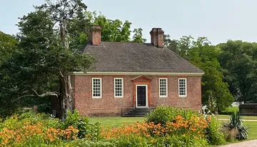 The image shows a traditional brick house with a gabled roof, two chimneys, and surrounded by lush gardens.