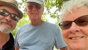 Three smiling people wearing sunglasses and hats are posing for a close-up selfie outdoors.