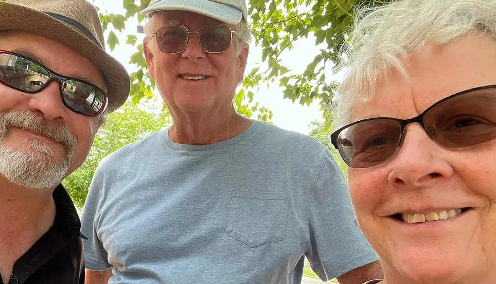 Three smiling people wearing sunglasses and hats are posing for a close-up selfie outdoors