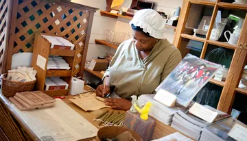 A person dressed in historical attire is working at a desk with documents and various old-fashioned items around.