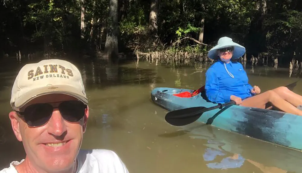A man in a cap is taking a selfie with a woman in a blue shirt and hat sitting in a kayak on a river surrounded by trees