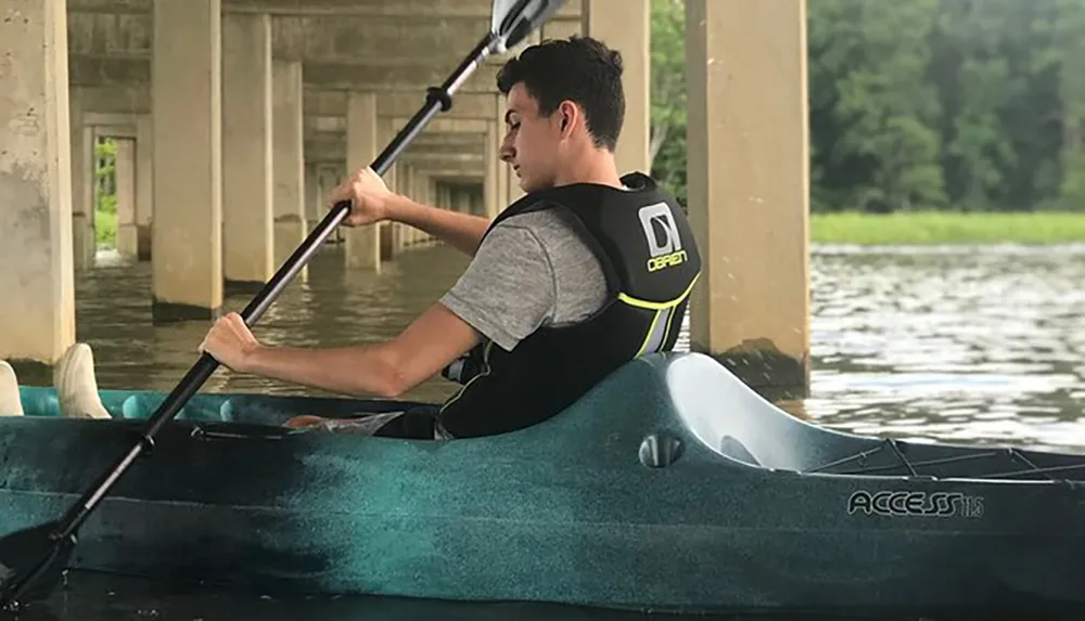 A person is kayaking under a bridge on a calm body of water wearing a life vest and actively paddling