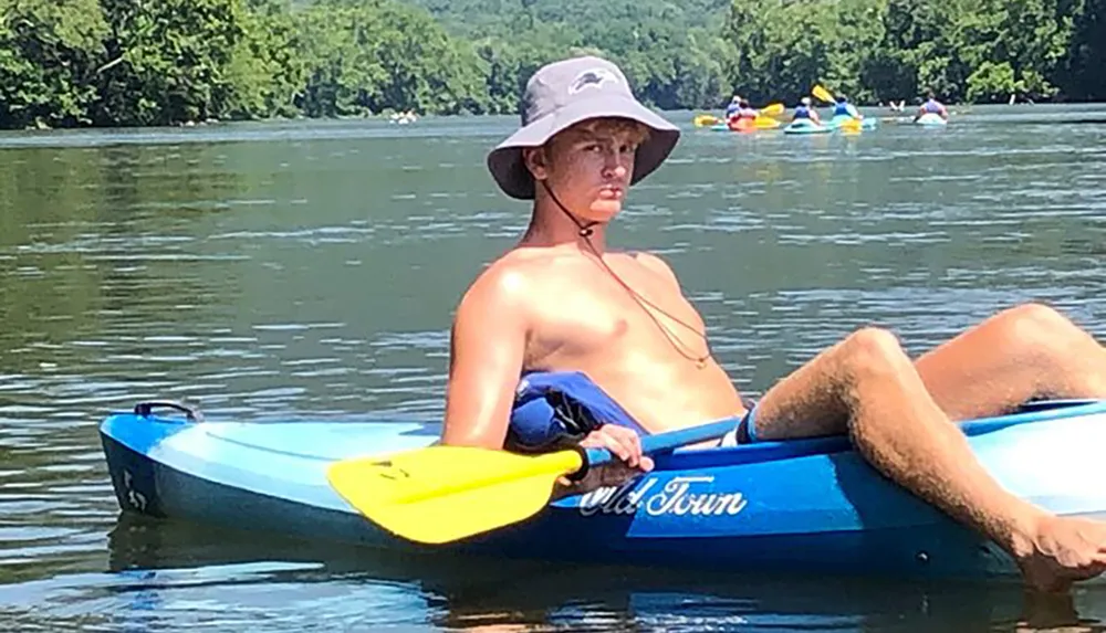A person lounges on a blue kayak in calm waters wearing a wide-brimmed hat and holding a yellow paddle