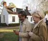 A smiling couple is looking at their smartphones while walking together on a sidewalk in front of a white historic-looking house