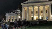 A group of people is gathered in front of a neoclassical building with white columns at nighttime, illuminated by exterior lights.