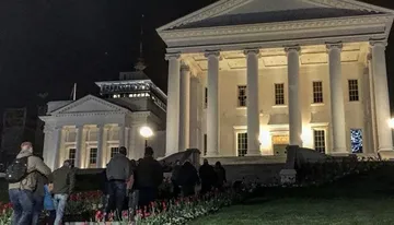 A group of people is gathered in front of a neoclassical building with white columns at nighttime, illuminated by exterior lights.