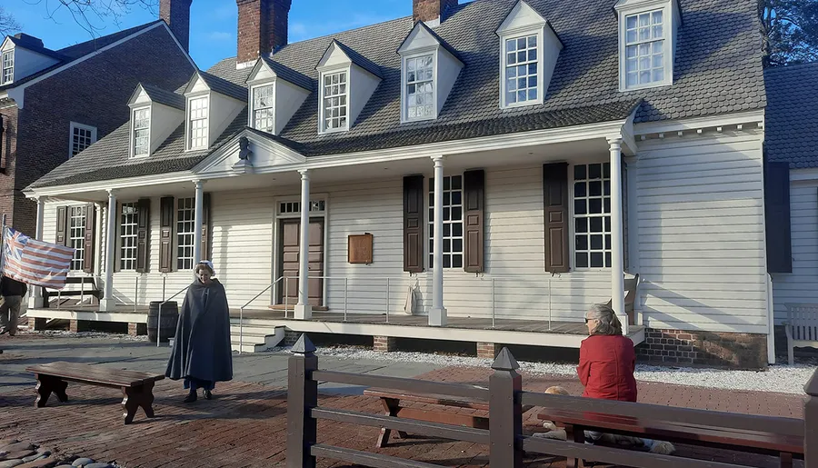 This image depicts a person in colonial attire standing in front of a historical building with a colonial-era American flag, while another person sits on a bench to the side.