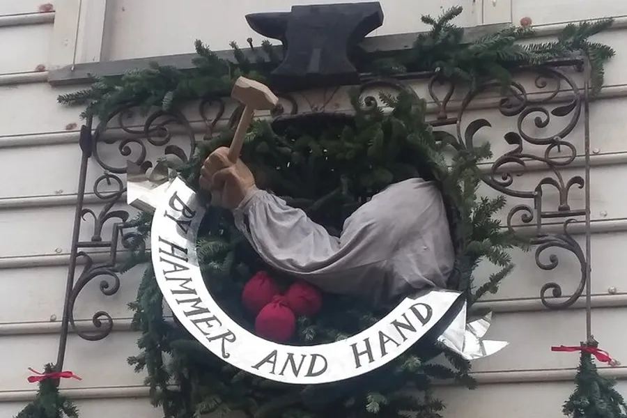 This image shows a three-dimensional emblem featuring an anvil and a hand with a hammer surrounded by a green wreath and a banner that reads HAMMER AND HAND.