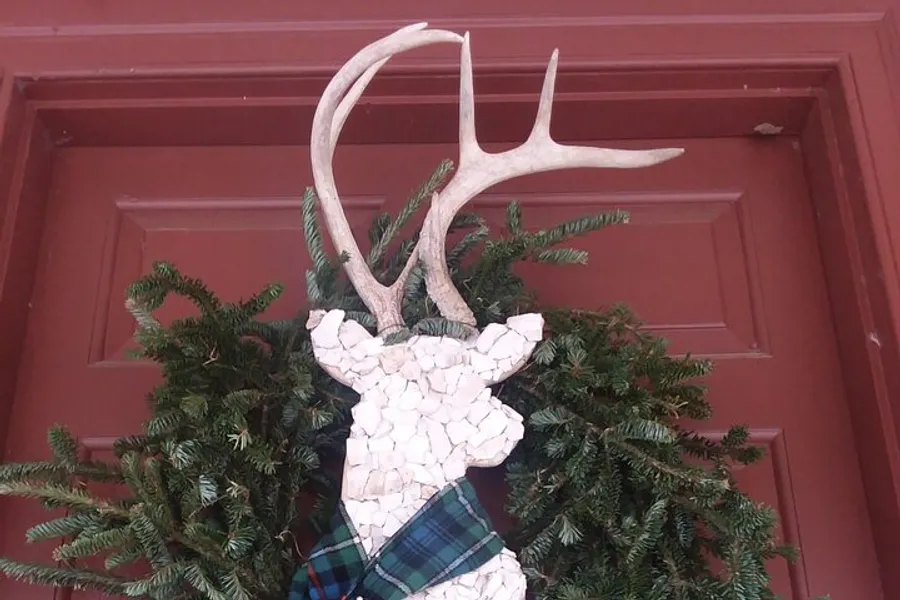 The image shows a decorative piece featuring a deer head silhouette with real antlers, pine branches, and a plaid ribbon against a red door.