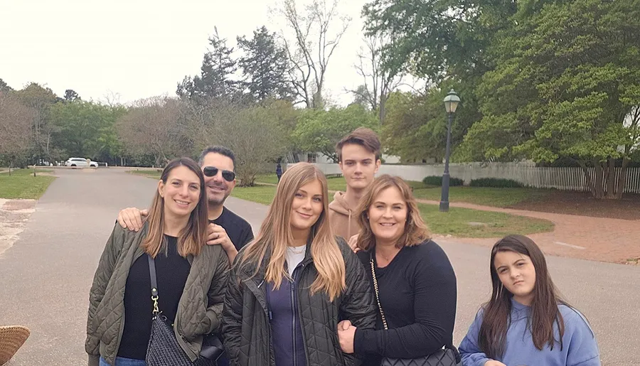 A group of six individuals, possibly a family, are smiling and posing for a photo outdoors on a path with trees and lampposts in the background.