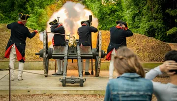 Individuals dressed in historical military uniforms are firing a cannon as spectators cover their ears.