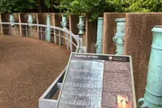 The image shows a curved walkway with a row of green cannons mounted on pedestals behind a metal barrier, with an information plaque in the foreground titled 
