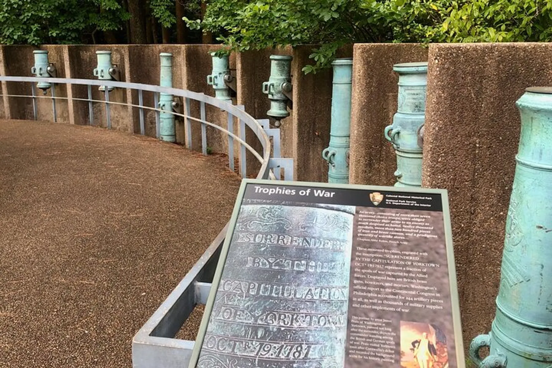 The image shows a curved walkway with a row of green cannons mounted on pedestals behind a metal barrier, with an information plaque in the foreground titled Trophies of War.