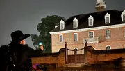 A person in a wide-brimmed hat is taking a reading with a handheld device outside a large brick building at night.