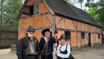 Three people dressed in historical costumes stand in front of a traditional half-timbered building, possibly at a historical reenactment site.