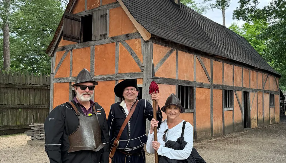 Three people dressed in historical costumes stand in front of a traditional half-timbered building possibly at a historical reenactment site