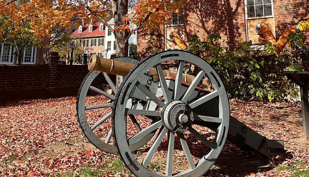 An old-fashioned cannon rests on a carriage amid fallen autumn leaves in a historical setting with colonial-style buildings in the background
