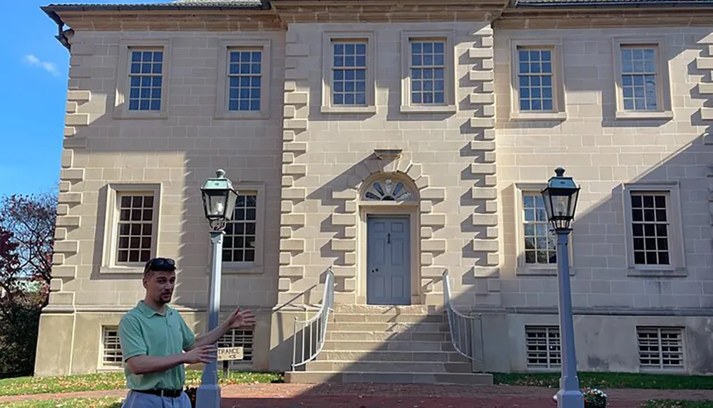 A person is gesturing with their hands while standing in front of a classical building with distinctive quoins and a central doorway