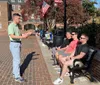 A person is gesturing while speaking to a group of individuals sitting on a bench in a sunny outdoor public space with American flags in the background