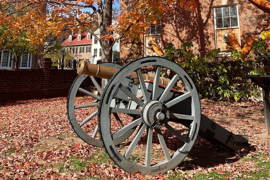 An old cannon on wooden wheels is displayed on a carpet of autumn leaves in front of colonial-style buildings with trees showcasing seasonal foliage in the background.