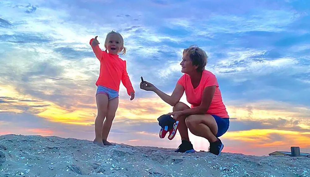 A young child excitedly points to the sky while an adult kneels beside her against a backdrop of a vibrant sunset at the beach