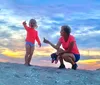 A child joyfully reaches out while a woman possibly her grandmother crouches beside her against a backdrop of a beautiful sunset on the beach