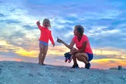 A child joyfully reaches out while a woman, possibly her grandmother, crouches beside her against a backdrop of a beautiful sunset on the beach.