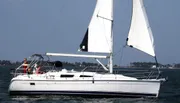 A sailboat with its sails unfurled glides through the water with a couple of people aboard.