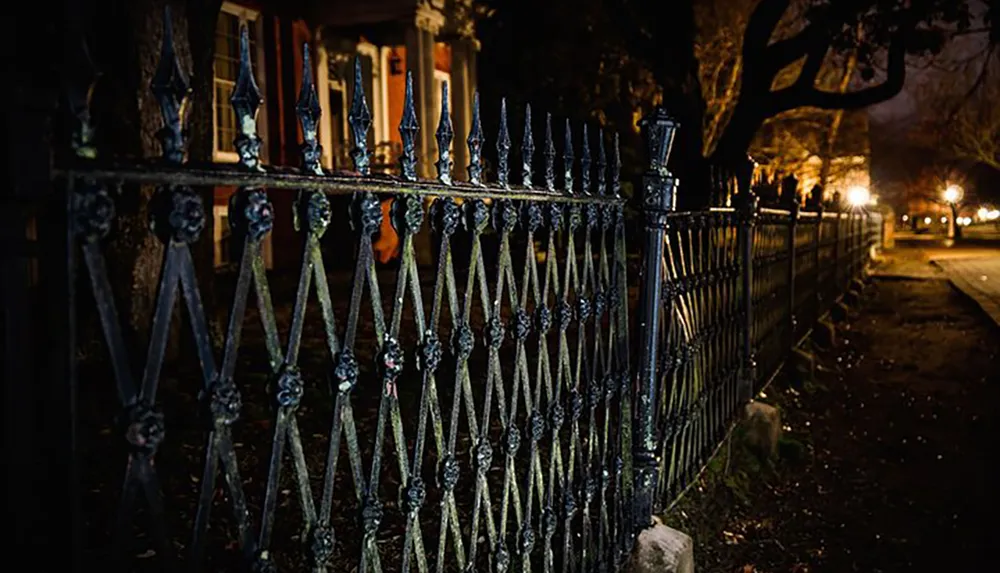 An ornate black iron fence stands in the foreground at night with warmly lit sidewalks receding into the background