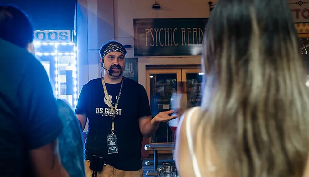 A man wearing a bandana and a US Ghost Adventures shirt is speaking and gesturing to a person with his back to the camera with a Psychic Readings sign in the background