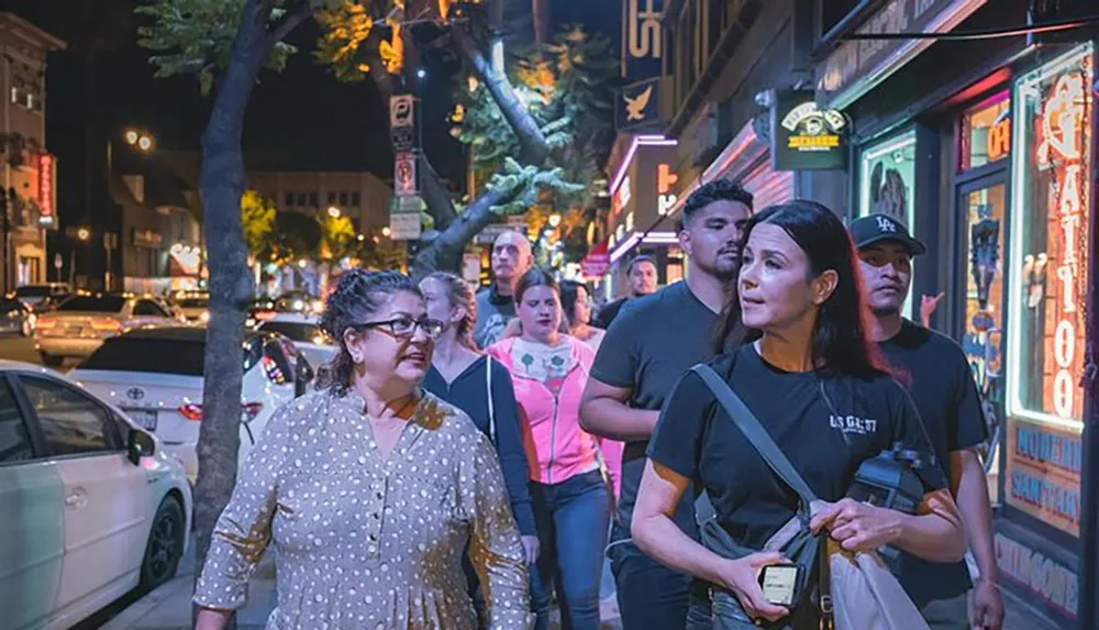 A group of people are walking on a city sidewalk at night passing by brightly lit shops and signs