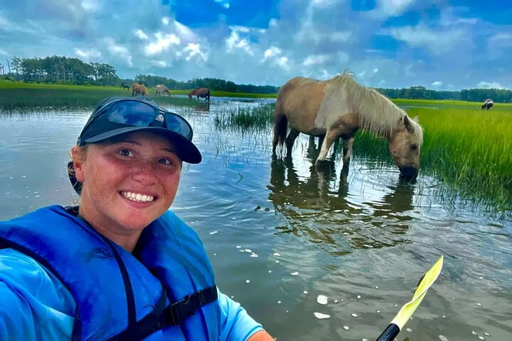 A person wearing a lifejacket is smiling for a selfie while kayaking near grazing horses in a scenic waterway with grass and trees in the background