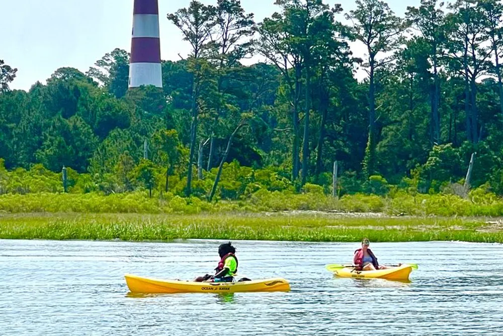 Two individuals are kayaking on a calm waterway with a tall lighthouse in the background amidst greenery