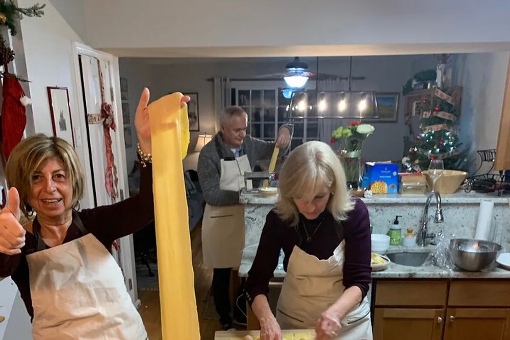 A person is proudly holding up a long sheet of pasta with a thumbs-up in a kitchen where others are also engaged in cooking activities