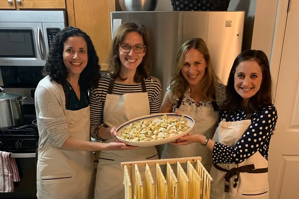 Four people are smiling for a photo in a kitchen proudly displaying a tray of homemade pasta with more pasta hanging to dry in the foreground